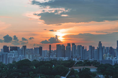 Buildings in city against sky during sunset with sun behind the cloud and park in foreground