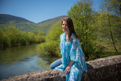Young woman sitting by lake against trees