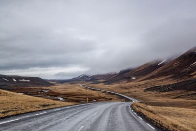 Country road leading towards mountains against cloudy sky