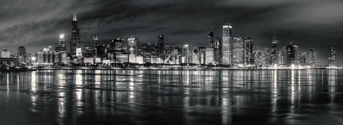 Illuminated modern buildings by river against sky at night