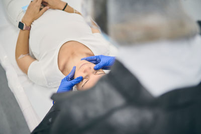 From above of beautician in gloves doing face massage for relaxed female client lying on table in beauty clinic