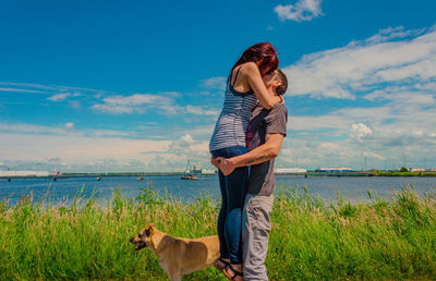 Man and woman kissing by dog on field against sky during sunny day