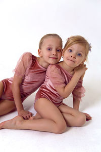 Two sisters sitting on a white background in pink bodysuits