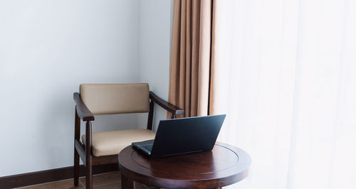 Black laptop on wood coffee table in bright room with empty chair, work space in hotel room.