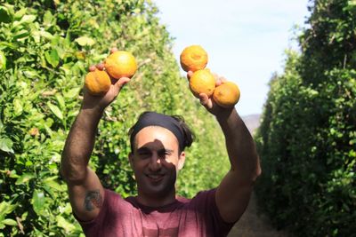 Portrait of man holding fruits by trees against sky