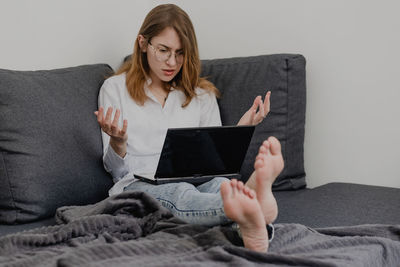 Attractive young woman working from home - female entrepreneur sitting on sofa with laptop computer and checking cell phone from comfort of home