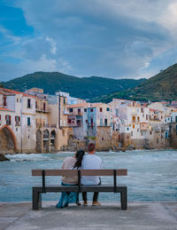 Rear view of people sitting on bench in city against sky