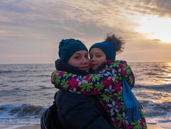 Mother and daughter in warm clothes on empty beach with blue sea on sunset