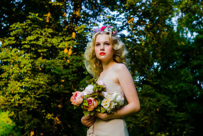 Beautiful bride holding flowers while standing against trees during wedding