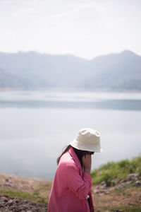 Side view of woman in hat by lake against sky