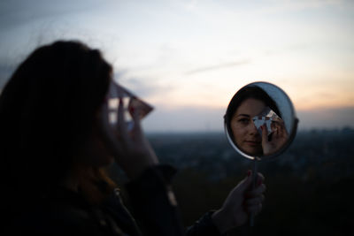 Reflection of woman holding prism in mirror against sky during sunset