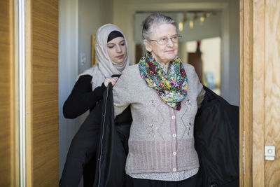 Young female home caregiver assisting senior woman in wearing jacket at home