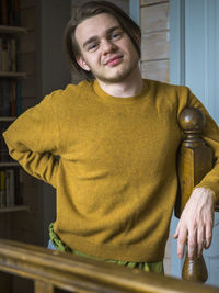 Portrait of smiling young man wearing sweater standing by wooden railing at home