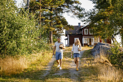 Full length of couple holding laptops while walking on footpath during sunny day