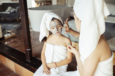 Mother in towel applying facial cream on daughter face while sitting at home