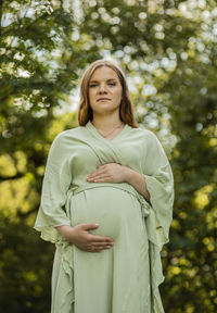 Young pregnant woman puts hands on big belly, green trees,blue sky on background. female wears dress