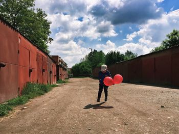 Playful boy with balloons on road against cloudy sky