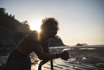 Shirtless young man looking away while leaning on parallel bars at beach