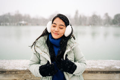 Happy young charming ethnic female in earmuffs and warm clothes feeling snowfall with closed eyes against city lake in madrid spain