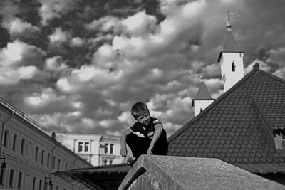 Boy crouching on railing in city against cloudy sky