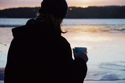 Rear view of woman holding drink at lakeshore during sunset