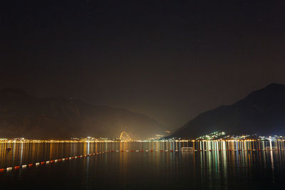 Illuminated city by lake against sky at night