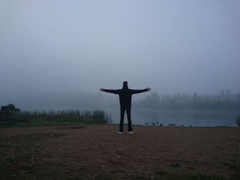 Rear view of man with arms outstretched standing by lake against sky in foggy weather