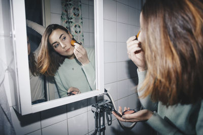 Young woman applying blush looking in mirror at college dorm bathroom