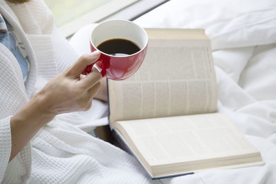 Midsection of woman with coffee cup and book on bed