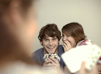 Woman whispering in young man's ear while playing cards with friend at home