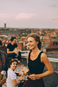 Smiling young female enjoying beer with friends at rooftop party