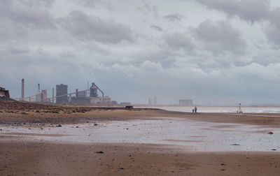 A woman and child walk along a seaside beach towards an industrial steel mill production plant.