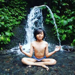 Shirtless boy meditating against waterfall