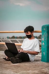 Young man using laptop wearing flu mask sitting by railing outdoors against sky