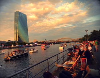 People on sea by city against sky during sunset