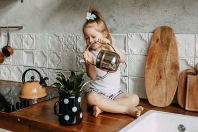 A little girl child takes care of watering a flower in a pot sitting on a kitchen cabinet at home