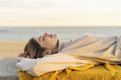 Young woman relaxing while lying on bench at beach promenade during sunset