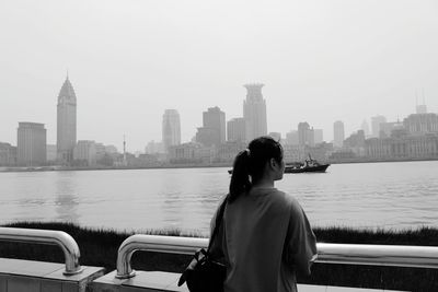 Rear view of man looking at river by city buildings against clear sky