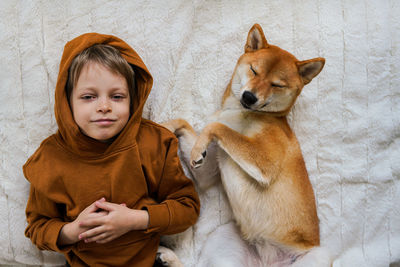 View from above on cute smiling boy and his domestic ginger dog are lying on white blanket