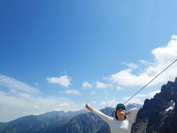 Low angle view of woman with arm outstretched against mountain ranges and sky during sunny day