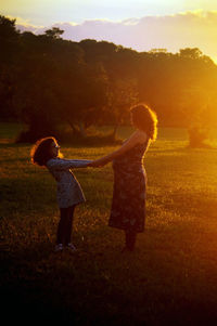 Girl and pregnant mother holding hands on field during sunset