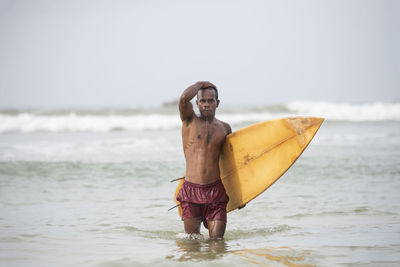 Portrait of shirtless man with surfboard wading in sea against sky