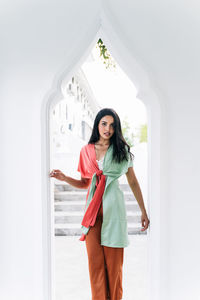 Cheerful charming young ethnic indian female in stylish clothes standing near on windowsill of arched window in oriental style house and looking at camera