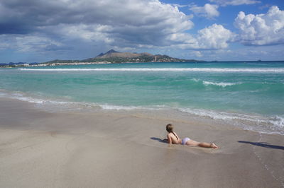 Full length of girl lying on shore at beach against cloudy sky
