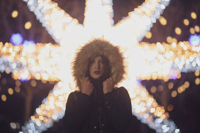 Portrait of woman wearing warm clothing against illuminated decoration at night