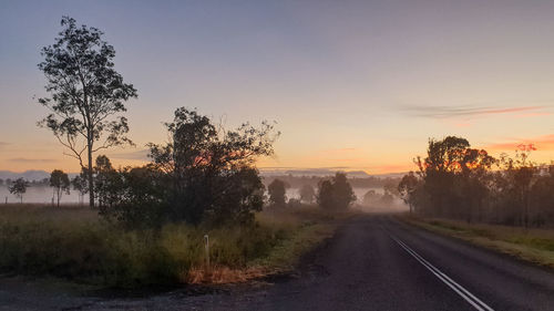 Road by trees against sky during sunset