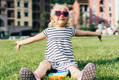 A funny little blonde in sunglasses and a striped dress sits on the lawn.