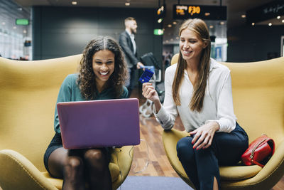 Smiling female multi-ethnic colleagues discussing over laptop and credit card while sitting at airport lobby
