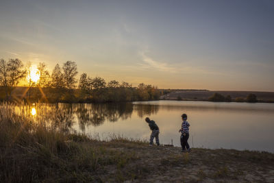 People standing by lake against sky during sunset