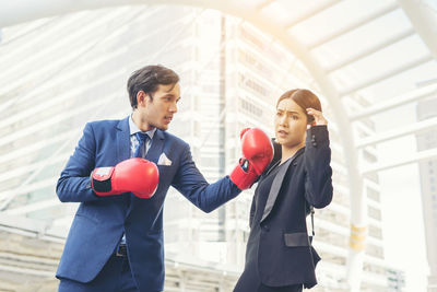 Young businessman hitting colleague with red boxing gloves against buildings in city